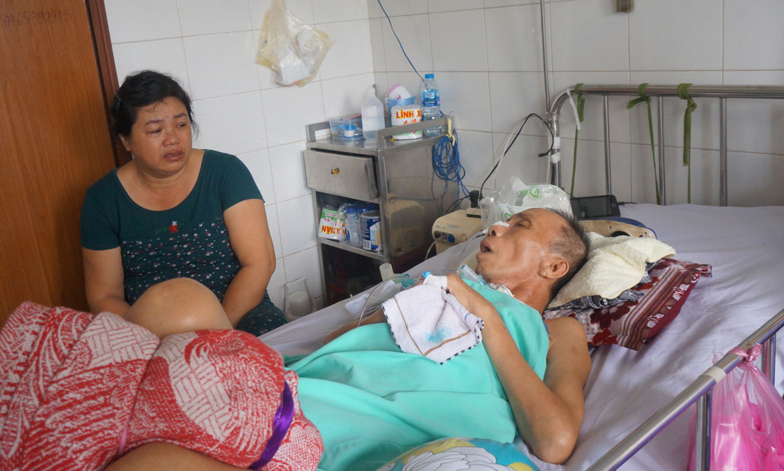 Bà Nguyễn Thị Chín khóc khi ngồi cạnh người chồng bị tai nạn giao thông chấn thương sọ não đang nằm điều trị hơn 5 tháng nay tại Bệnh viện Đa khoa Long An