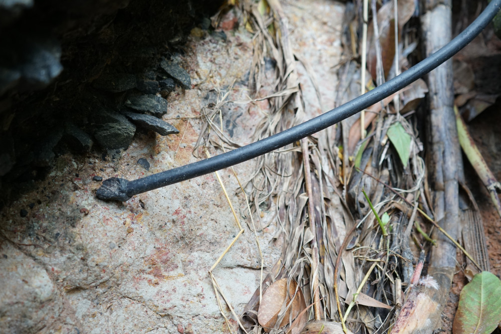 Một đoạn dây, trông giống như dây điện, bị đứt nằm dài theo mặt đường. Nếu là dây đang dẫn điện thì tiềm ẩn nguy cơ gây tai nạn điện cho người đi đường.